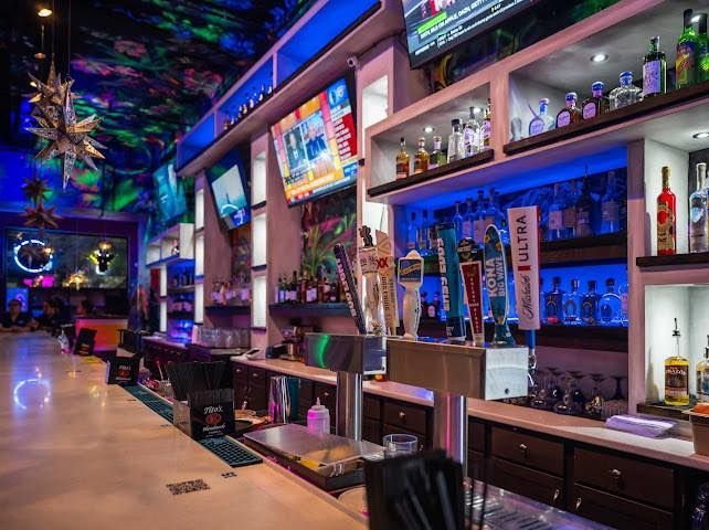 Bar interior with illuminated shelves, drinks, TVs, and a colorful, lit ceiling.