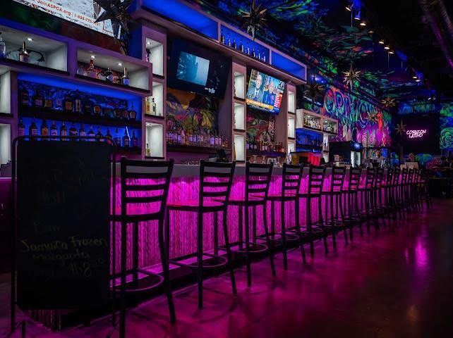 Bar interior with bright purple and blue lights. Tall bar stools line a backlit bar, shelves with bottles in the background.