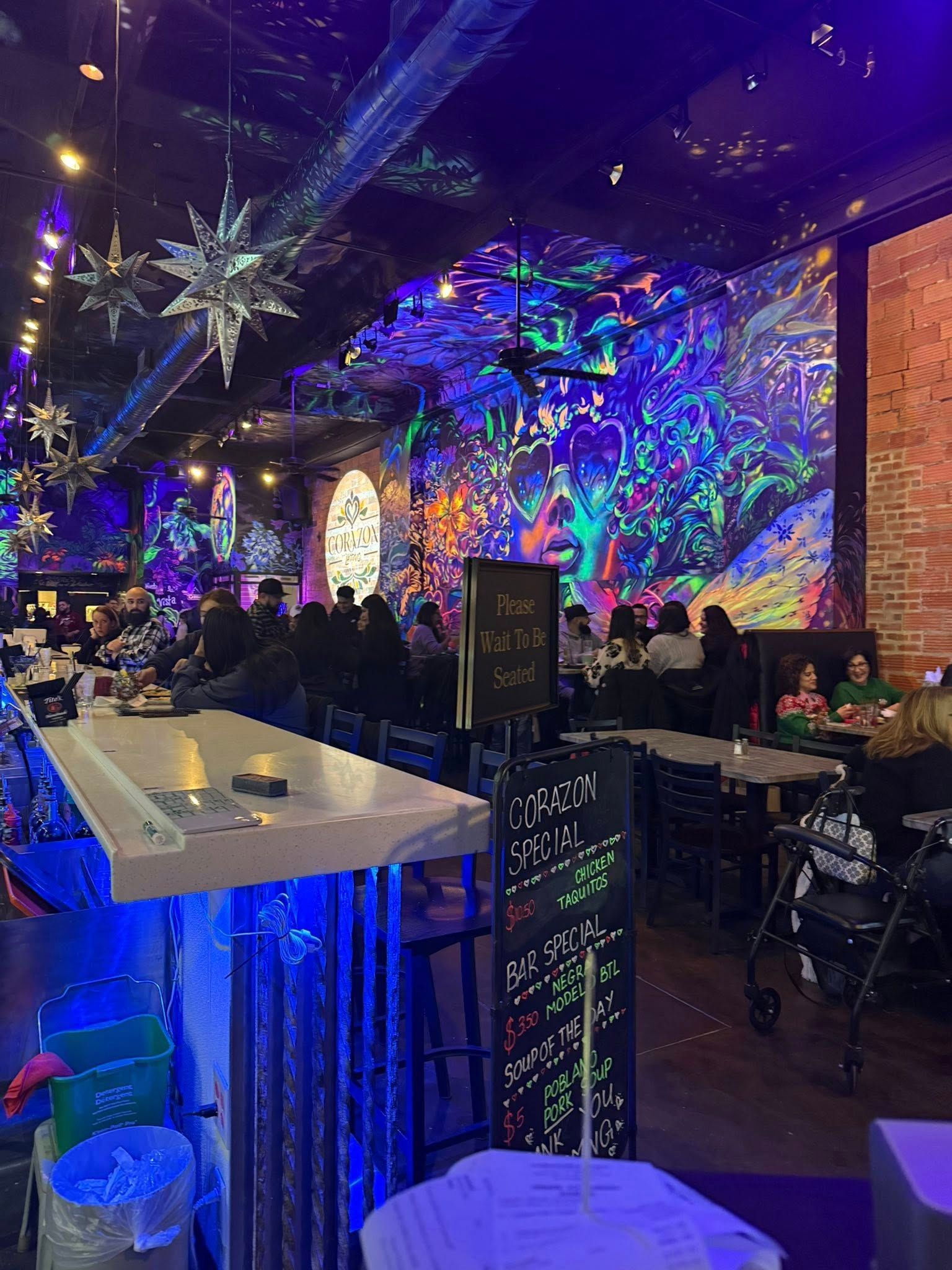Interior of a restaurant with blacklight art, patrons, bar, and tables. Bright colors and starry decorations.