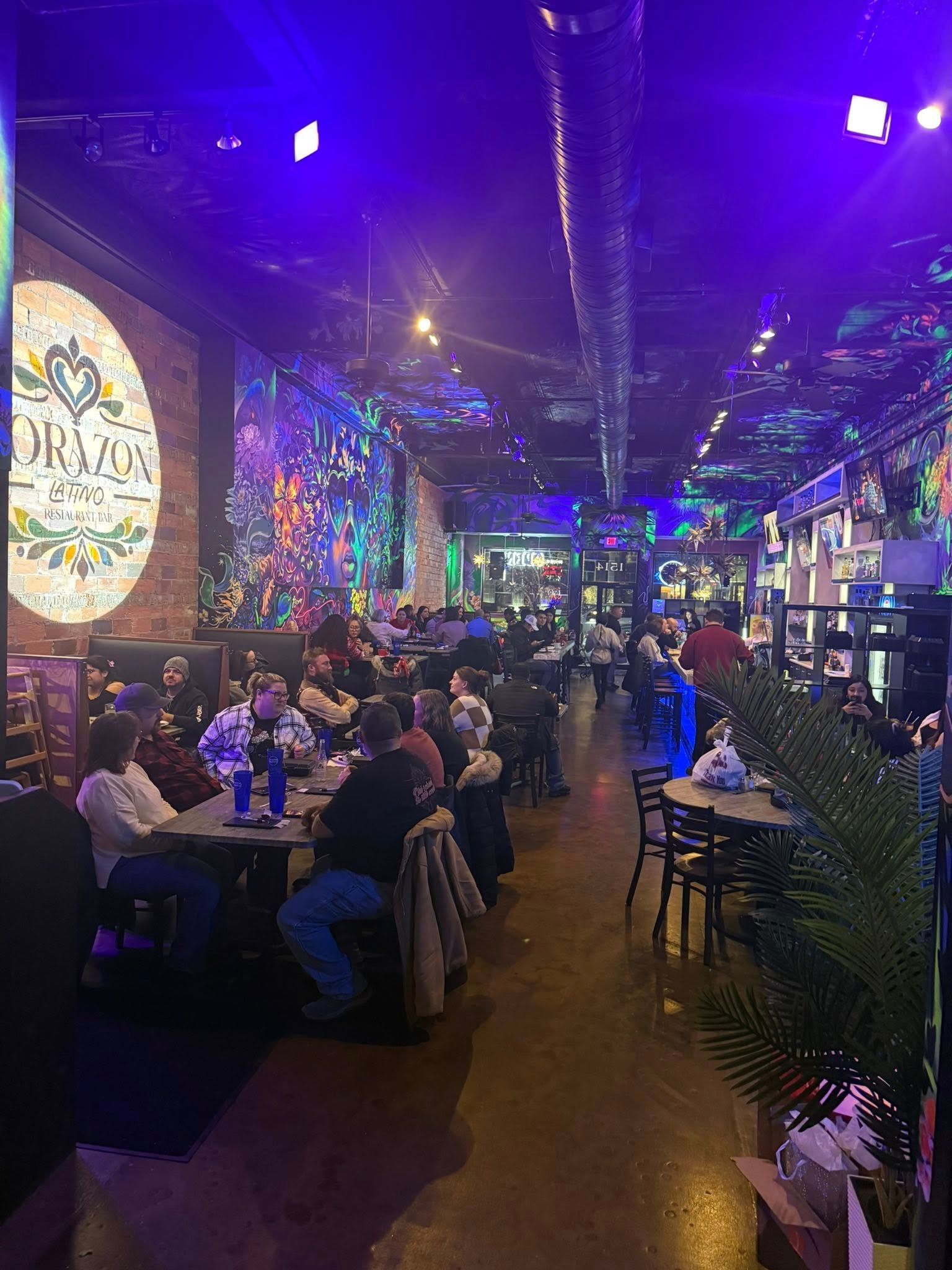 Interior of a dimly lit restaurant with patrons seated at tables.  Walls have colorful murals and there is a bar in the background.
