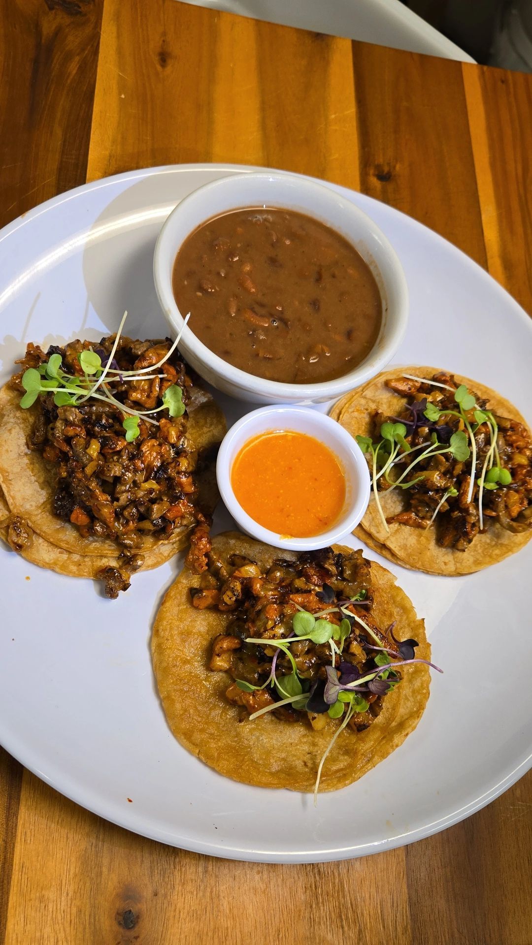 Tacos on a white plate with a side of sauce and refried beans. Microgreens top each taco.