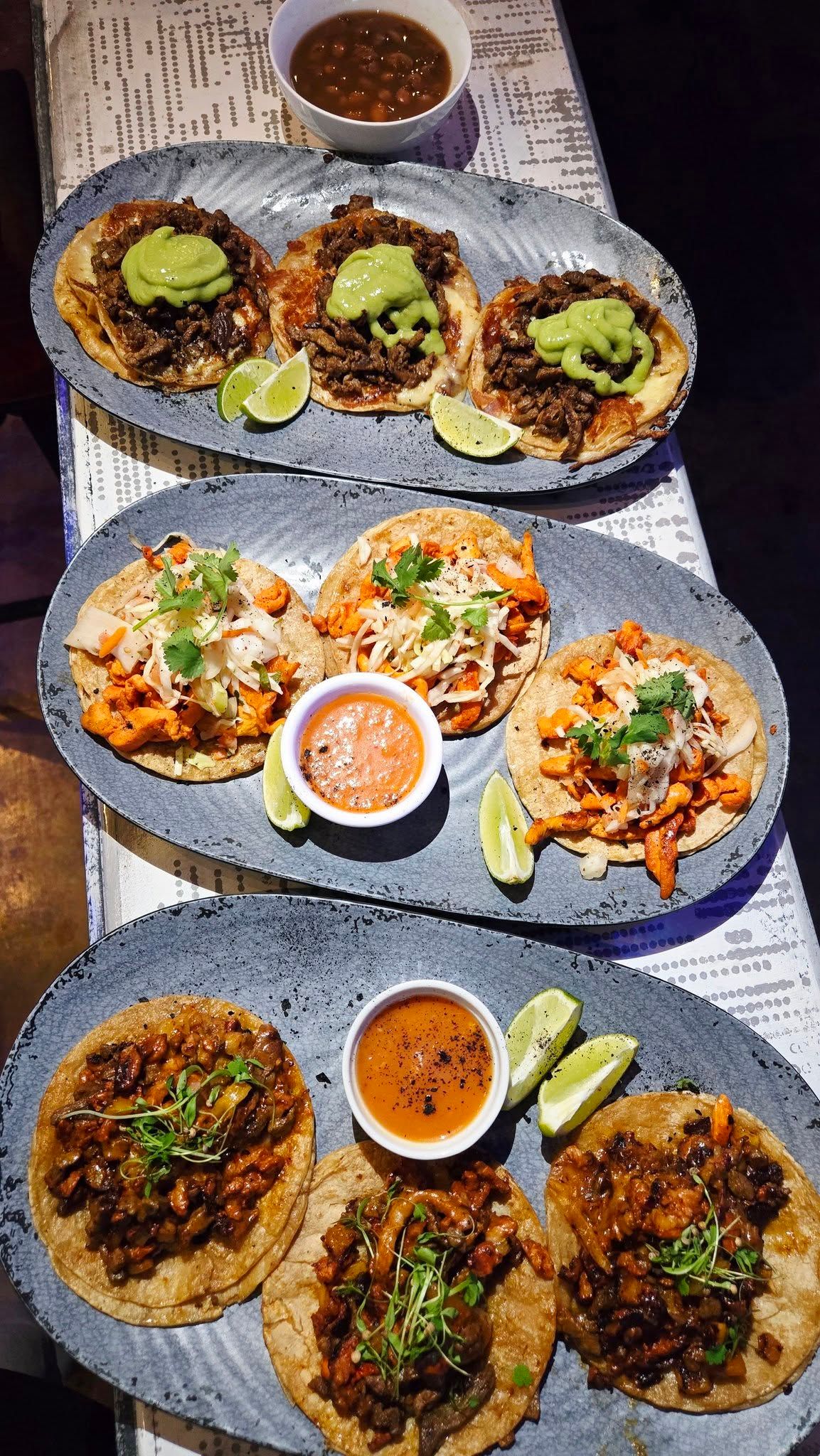 Three platters of tacos with various fillings, lime wedges, and dipping sauces.