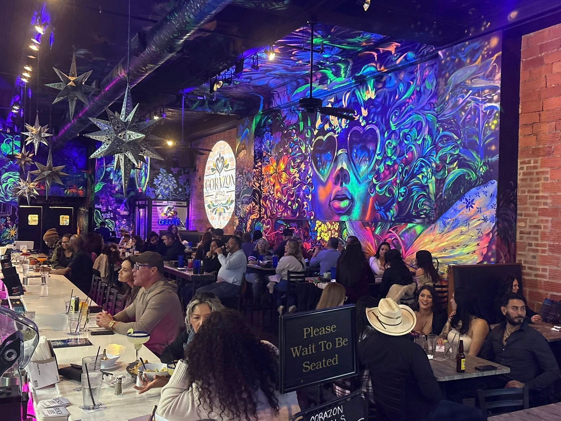 Interior of a crowded restaurant with a bar, colorful mural, and patrons at tables and bar.