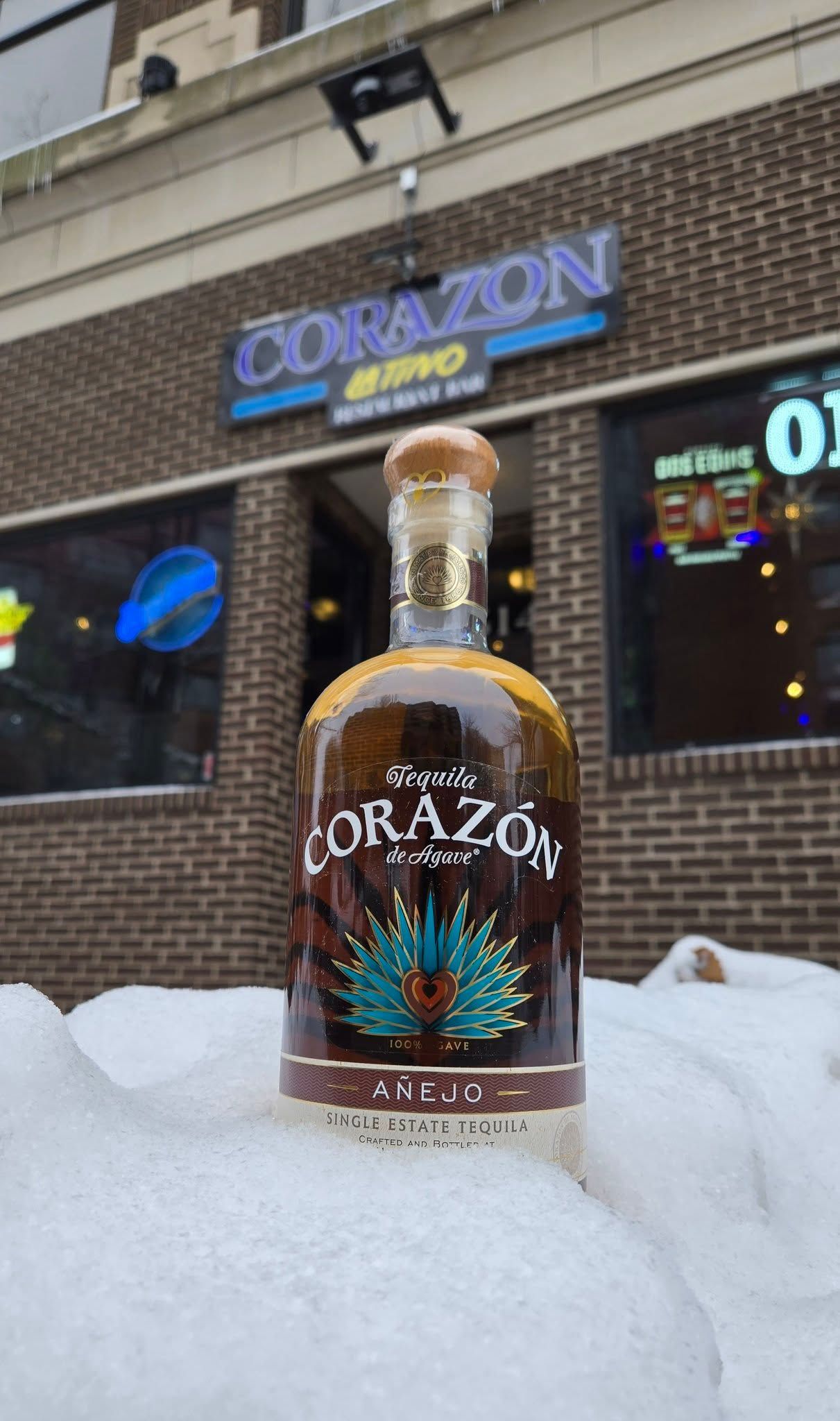 Bottle of Corazon tequila on snow outside a brick building with 
