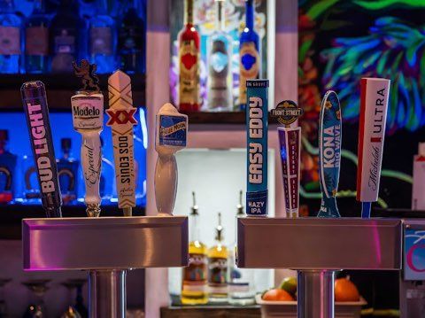 Beer taps at a bar with various brand handles. Backlit bottles, fruit and cocktails are in the background.