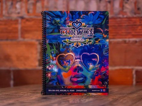 Corazon Mexican restaurant menu with vibrant artwork of a face wearing heart-shaped sunglasses, spiral-bound, set against a brick wall.