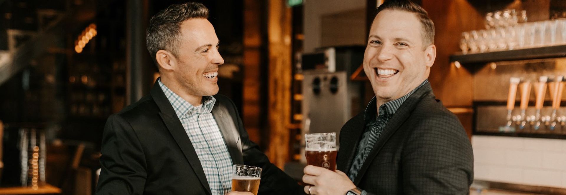 Two men are standing next to each other in a bar holding glasses of beer.