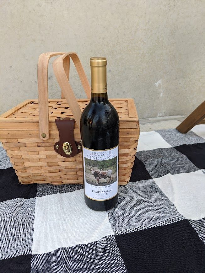 A bottle of wine is sitting next to a picnic basket on a checkered blanket.