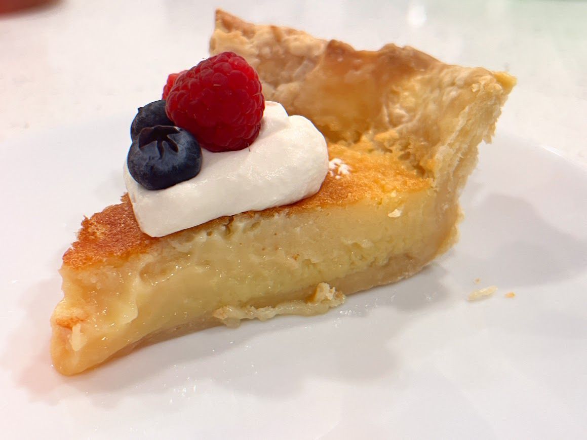 A slice of buttermilk pie with whipped cream and berries on top on a white plate.