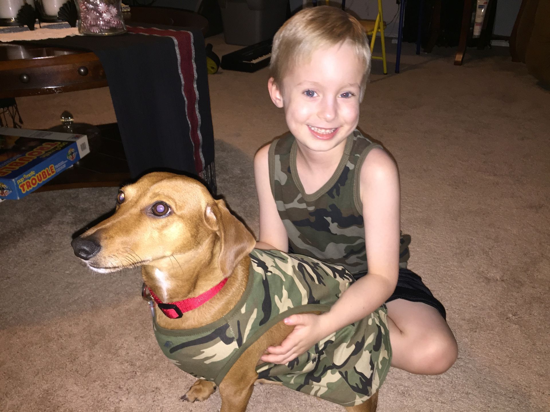 A young boy is kneeling next to a dog wearing a camouflage shirt