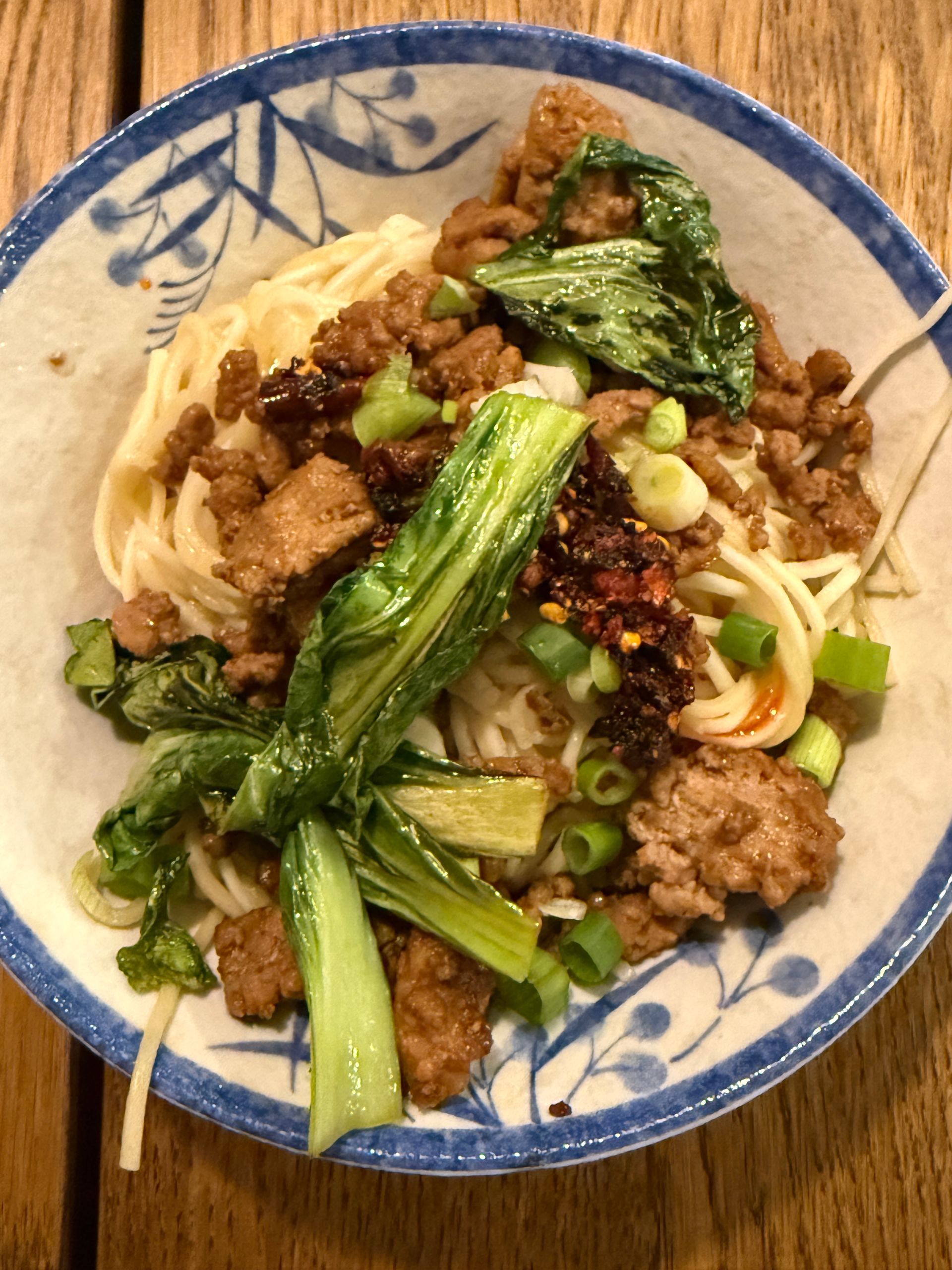 A bowl of noodles with meat and vegetables on a wooden table.