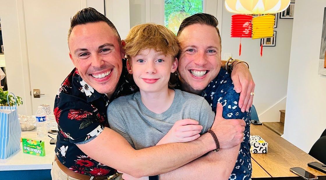 Three men are hugging a young boy in a living room.
