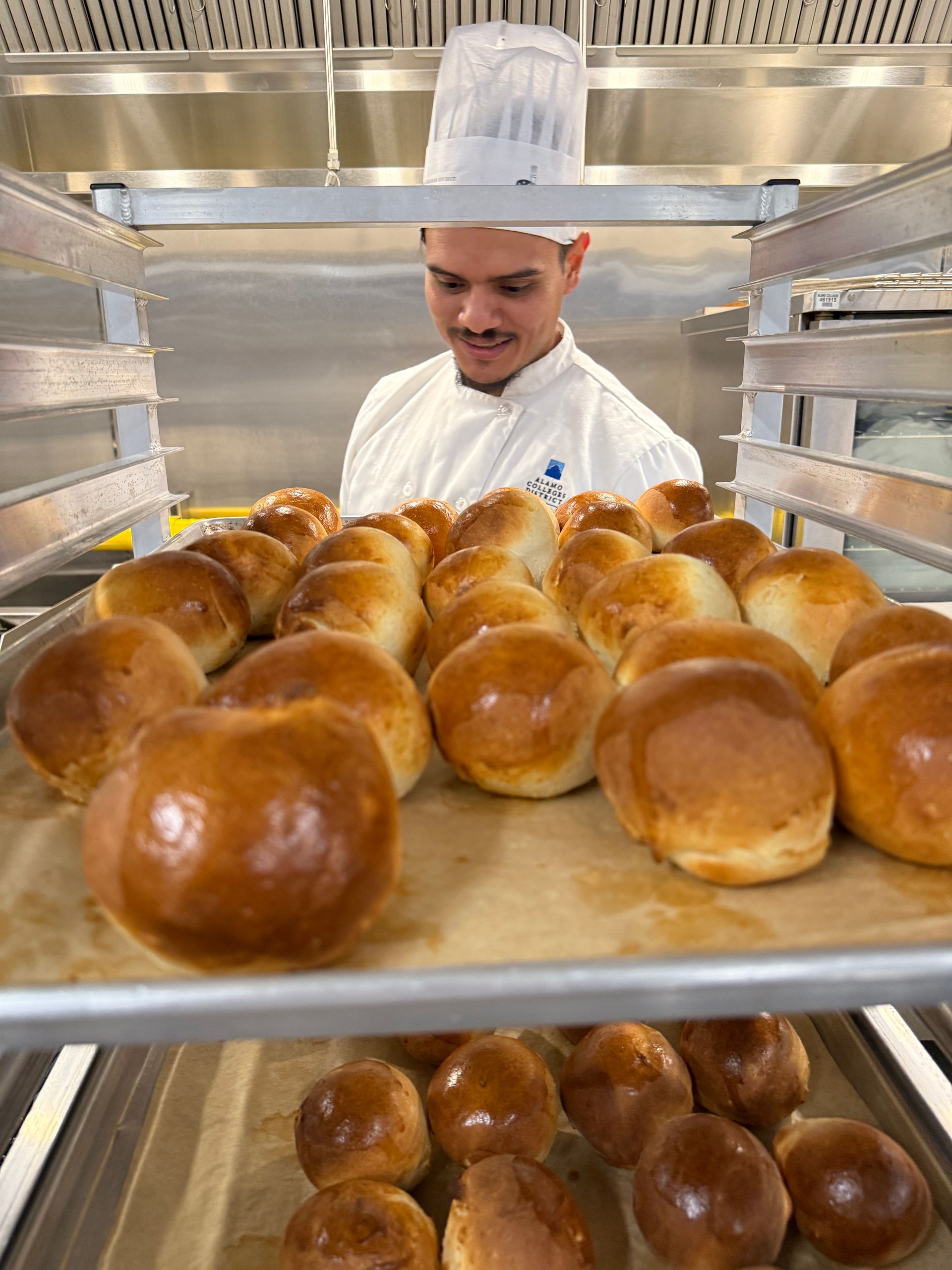 A man in a chef 's hat is standing in front of a tray of rolls.
