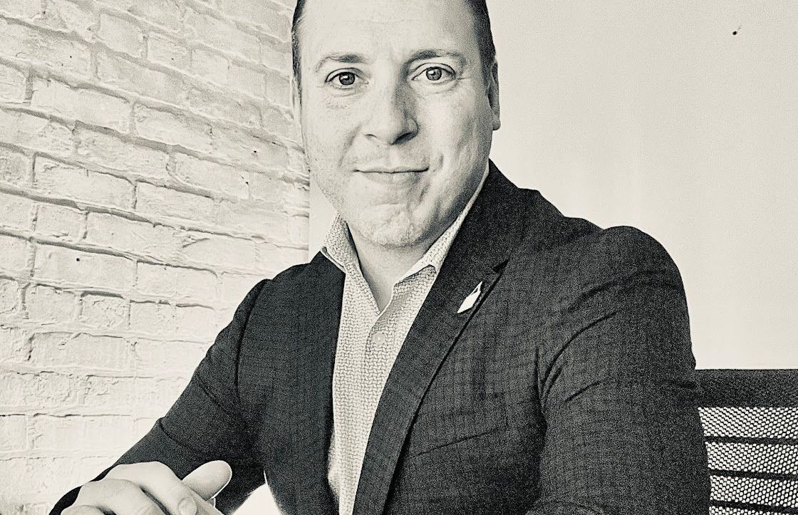 A man in a suit is sitting at a desk with his hands folded.