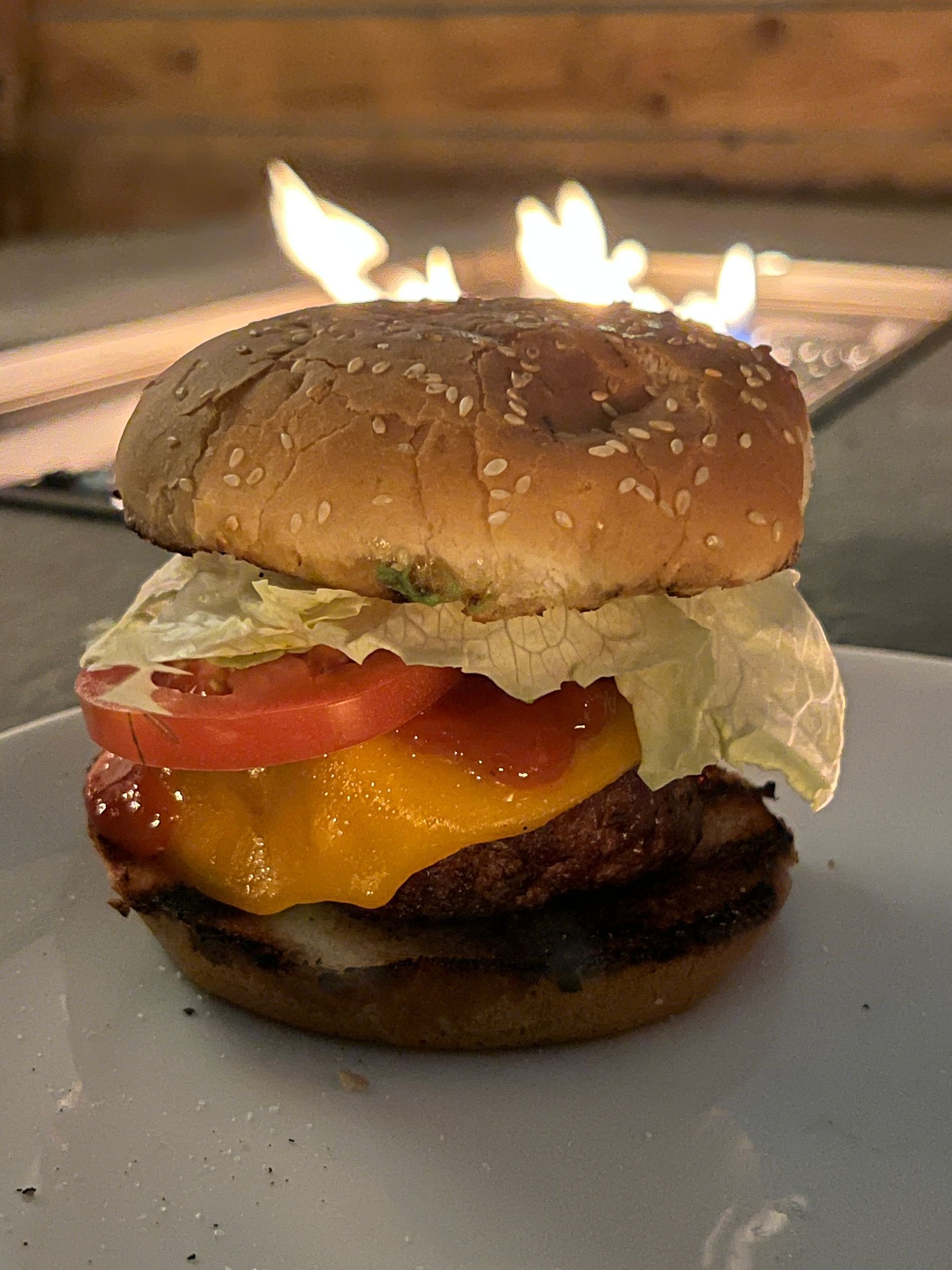 A close up of a hamburger on a plate with a fire pit in the background.