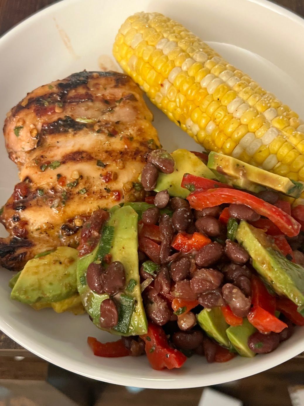 A white plate topped with chicken , beans , corn on the cob and avocado.