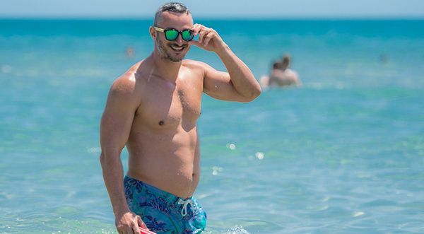 A shirtless man wearing sunglasses is standing in the ocean.