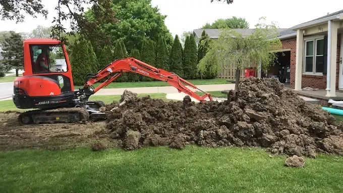 Small red excavator moving dirt pile beside a house and lawn