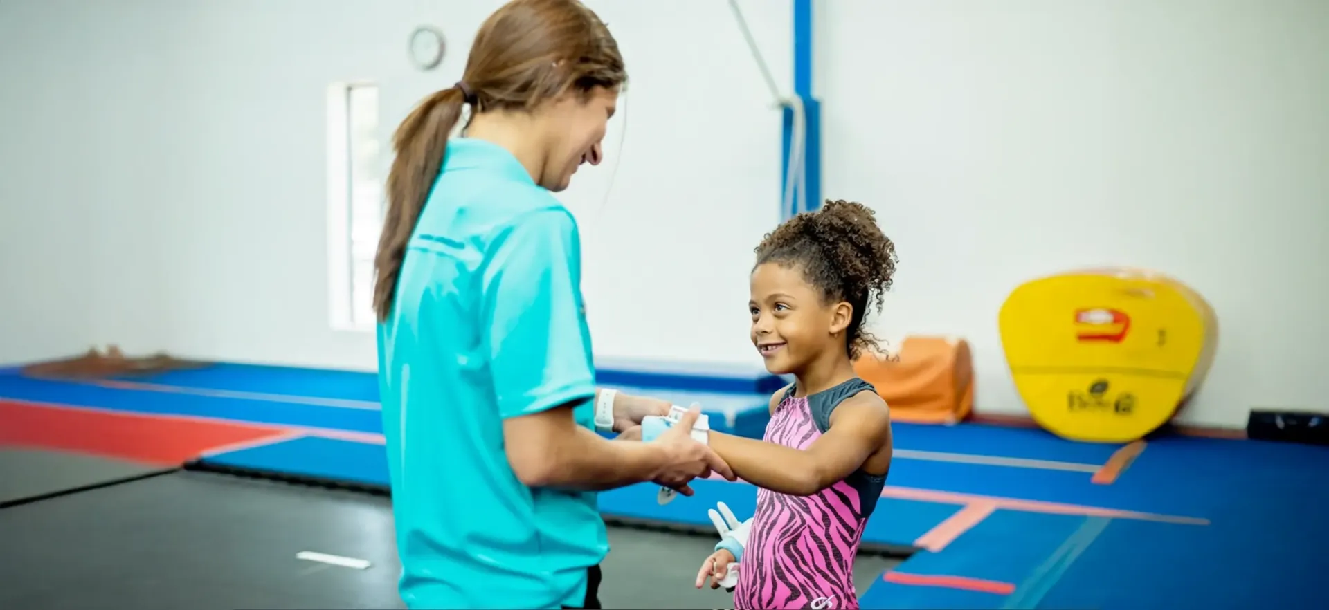 A coach assisting a student with their grips in a gym, with mats and a yellow barrel in the background