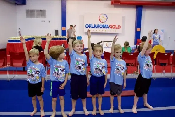 Children in blue shirts raise hands at Oklahoma Gold Gymnastics.
