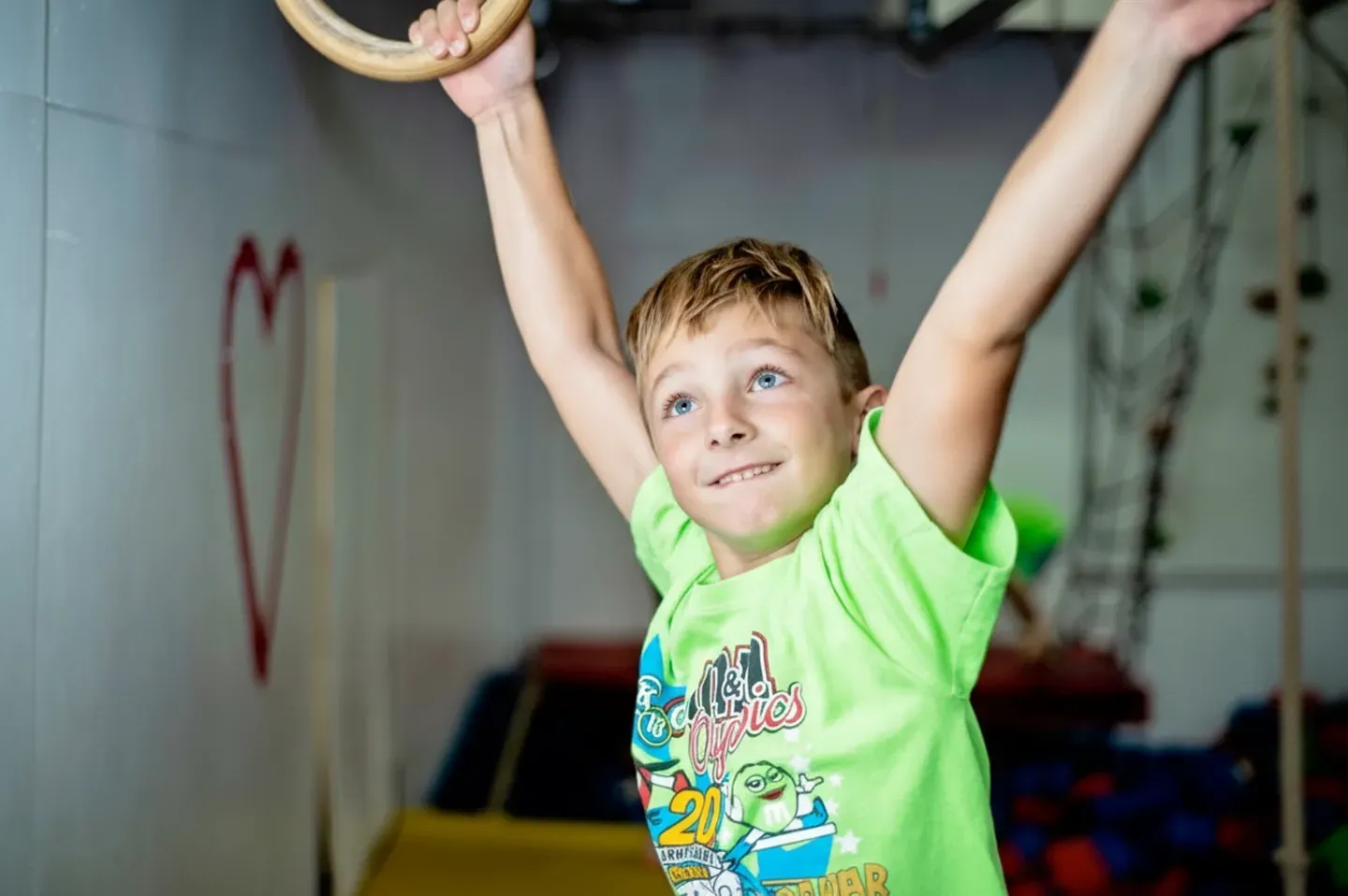 Boy hangs from gymnastic rings, smiling.  He wears a neon green shirt indoors.