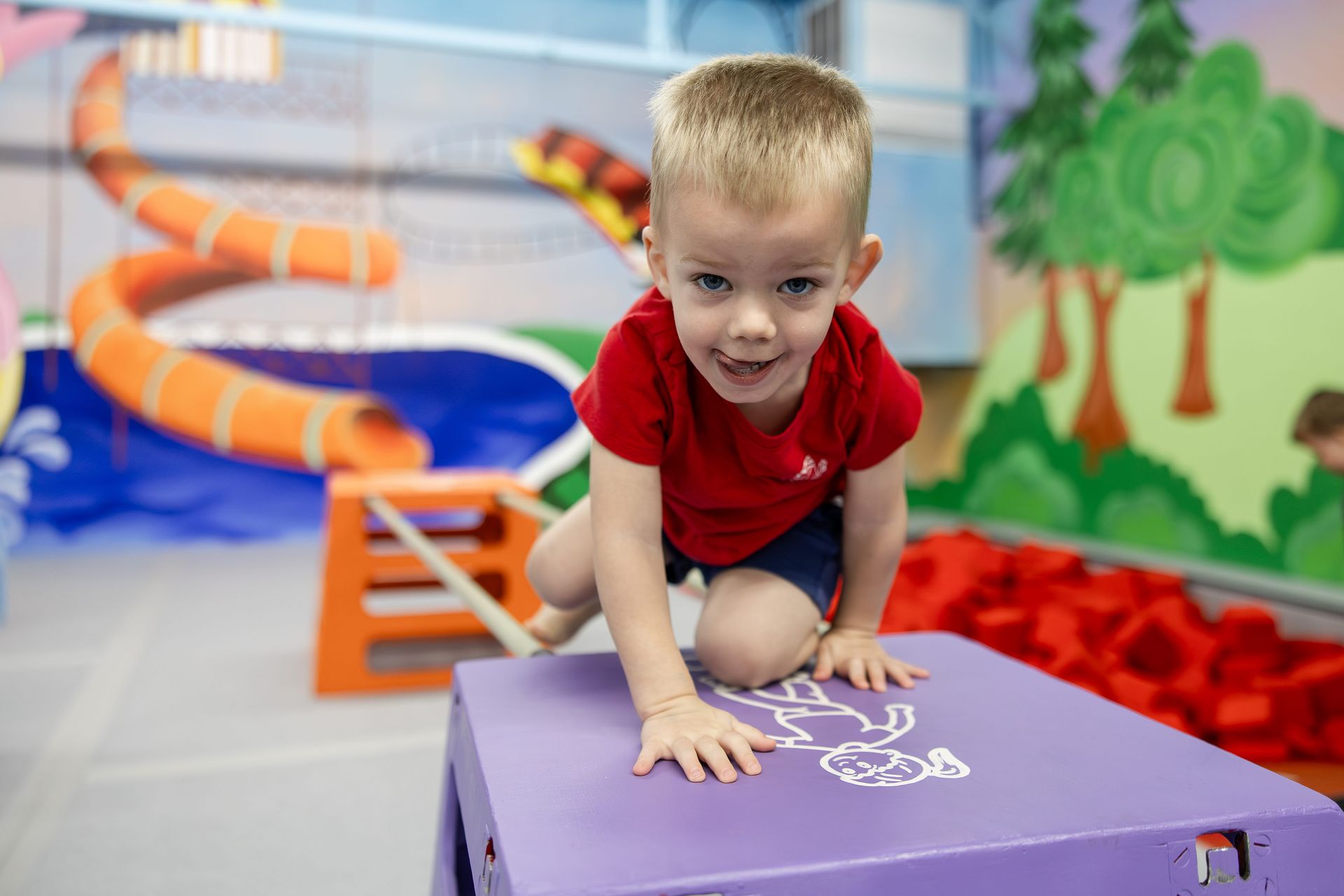 Toddler climbing on a purple padded block in an indoor play area, smiling at the camera.