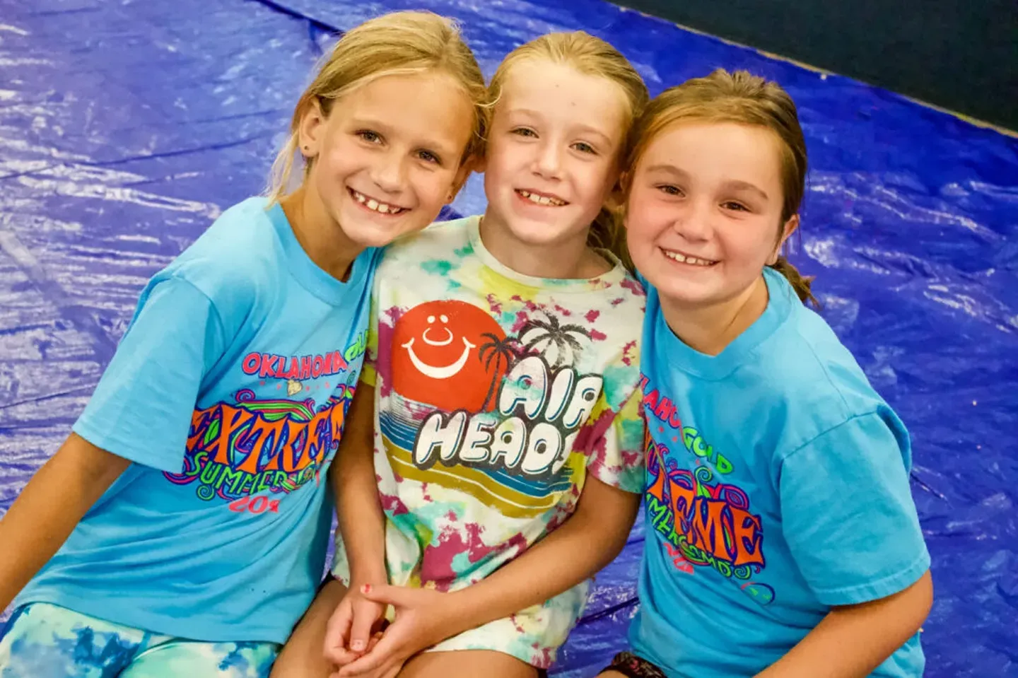 Three smiling girls in blue and tie-dye shirts, posing close together against a blue background.