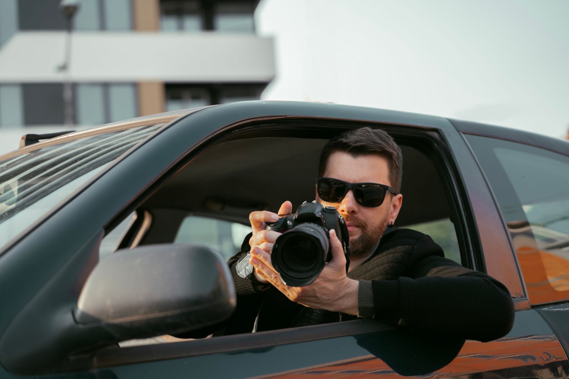 Person holding a camera and taking a photo from a car window near a modern building.