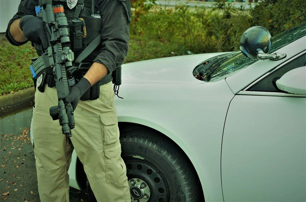 Police officer in tactical gear holding a rifle, next to a white patrol car.