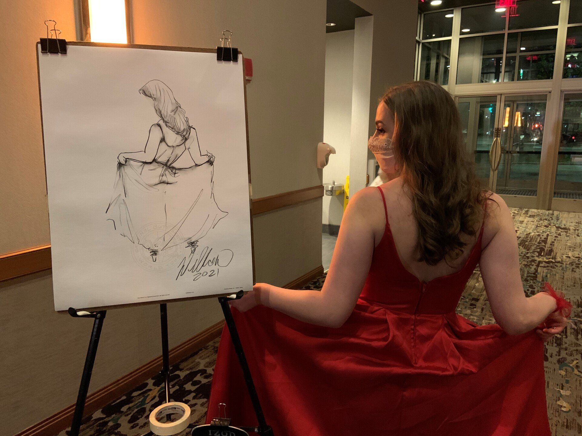 A woman in a red dress is sitting in front of an easel with a drawing on it.