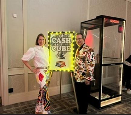 Two people standing beside a neon “CASH CUBE” game booth and a clear acrylic cube on wheels.
