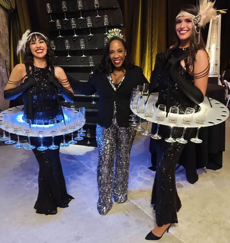 Three women in flapper attire, two with champagne trays, posing with a woman in a blazer and sequined pants.