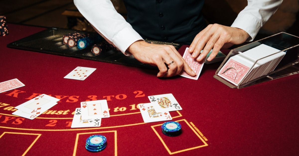A croupier in a white shirt and dark vest deals playing cards on a red blackjack table. It features chips and a card deck.