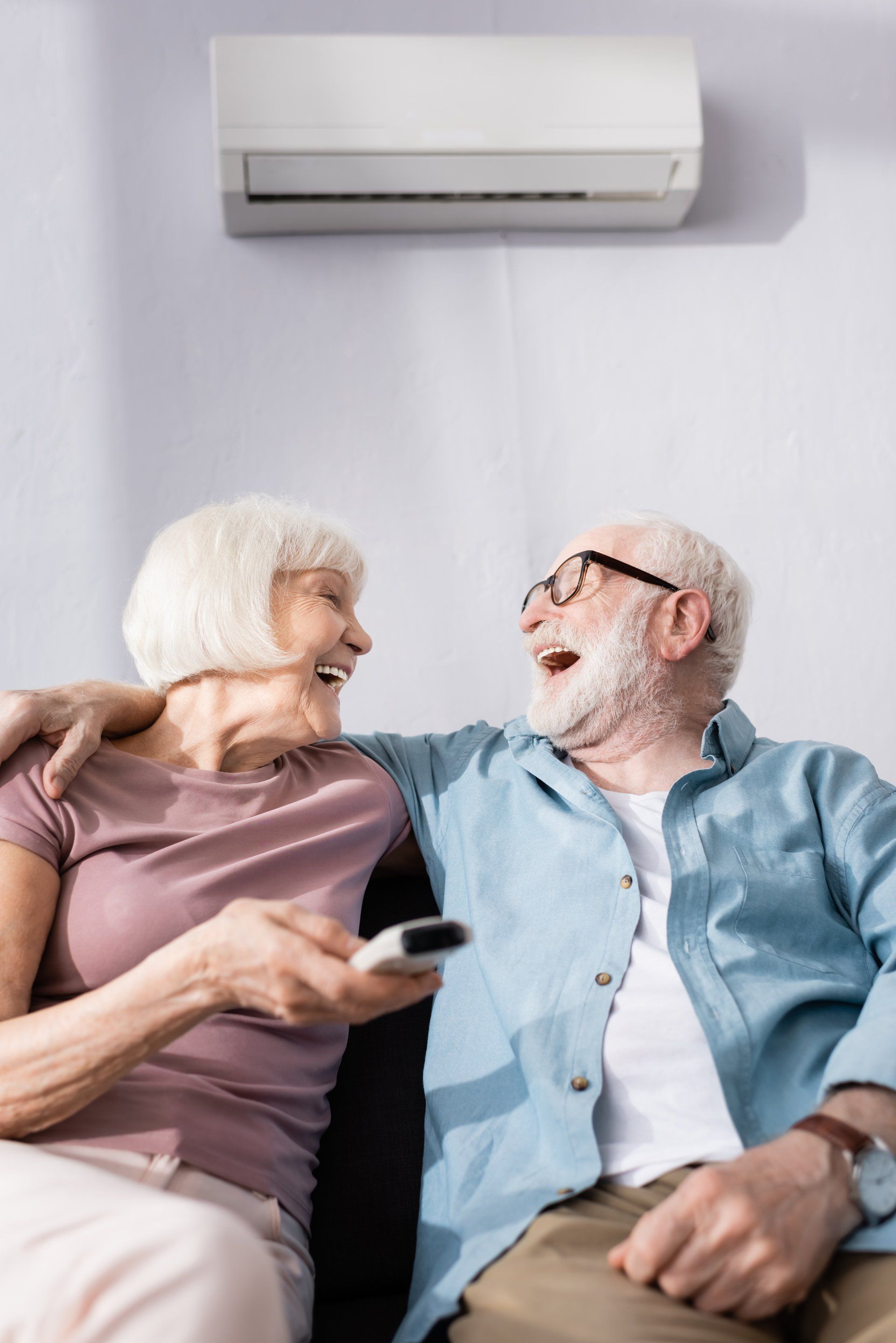 Selective focus of laughing senior man embracing wife with remote controller of air conditioner