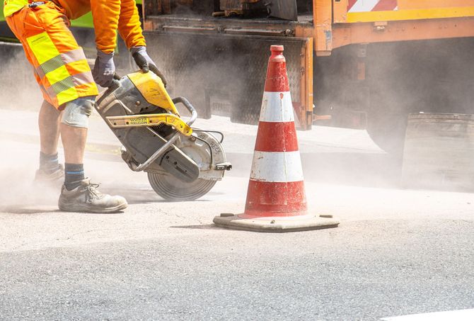 Worker cutting pavement beside an orange traffic cone, sending up dust at a roadwork site