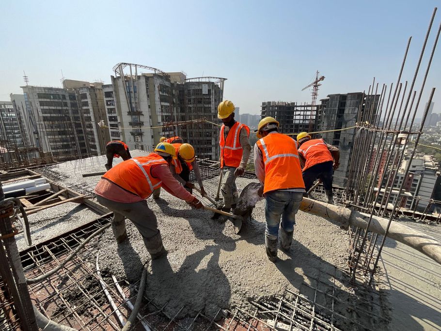 Construction workers in orange vests pour concrete on a rooftop with rebar and city buildings in the background