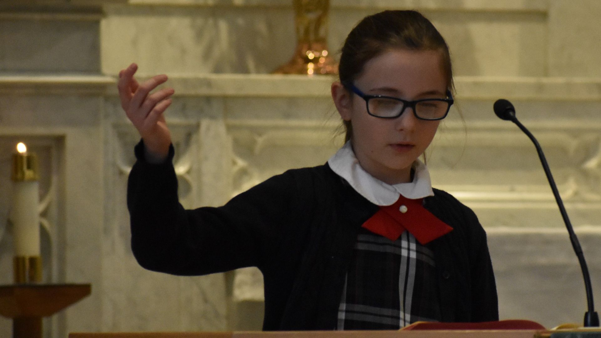 A young girl wearing glasses is giving a speech in front of a microphone