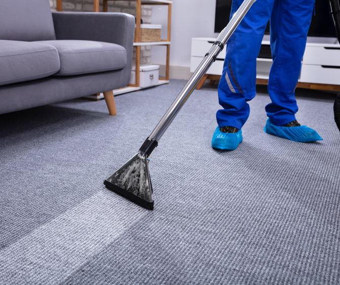 Person in blue coveralls cleans a gray carpet with a carpet cleaner in a living room.