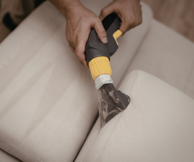 Person cleaning a light-colored couch with a handheld upholstery cleaner, spraying and extracting dirt.