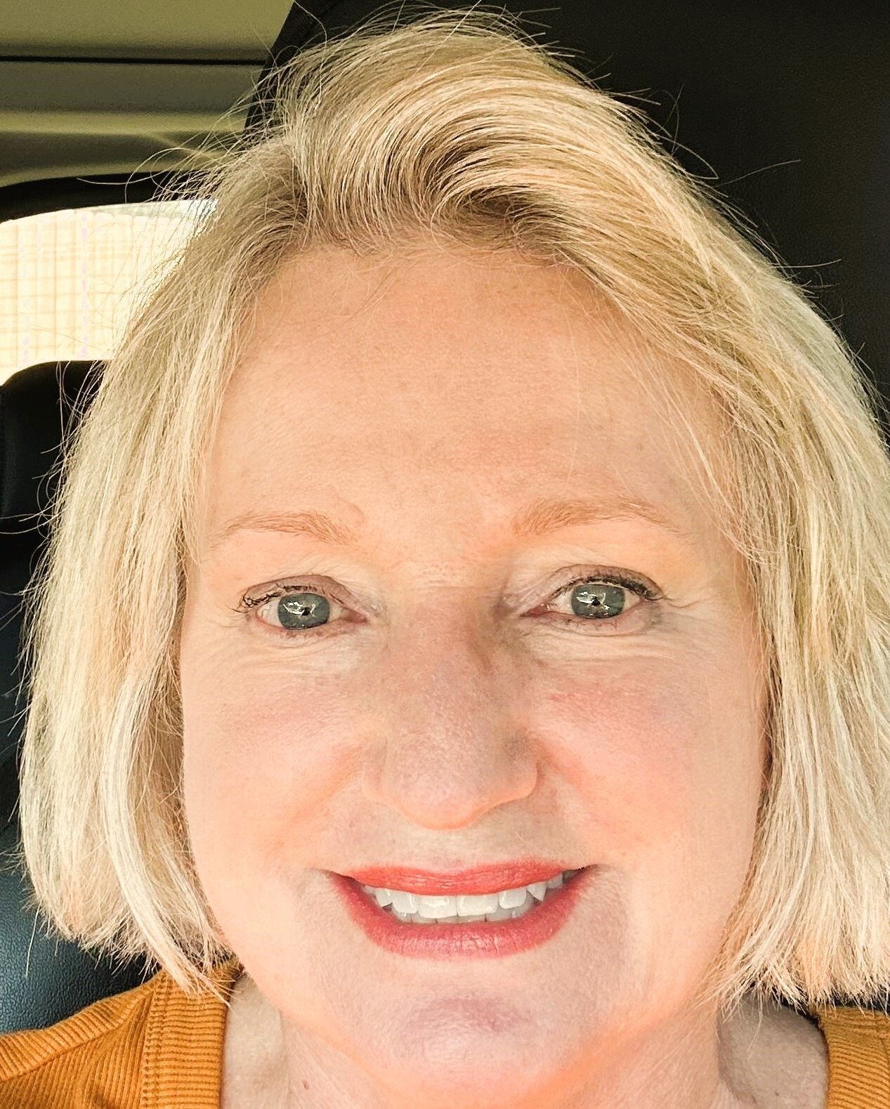 Blond woman smiling, wearing an orange sweater, set in front of a dark interior.