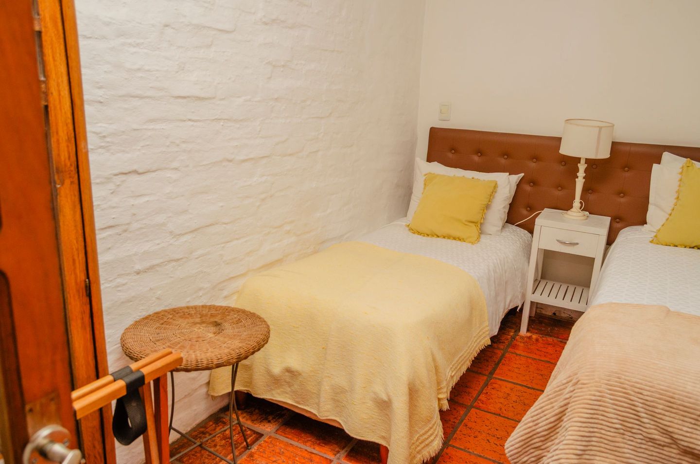 Un dormitorio con dos camas individuales, almohadas amarillas, una mesita de noche blanca con lámpara y suelo de baldosas junto a una pared de ladrillo blanco.