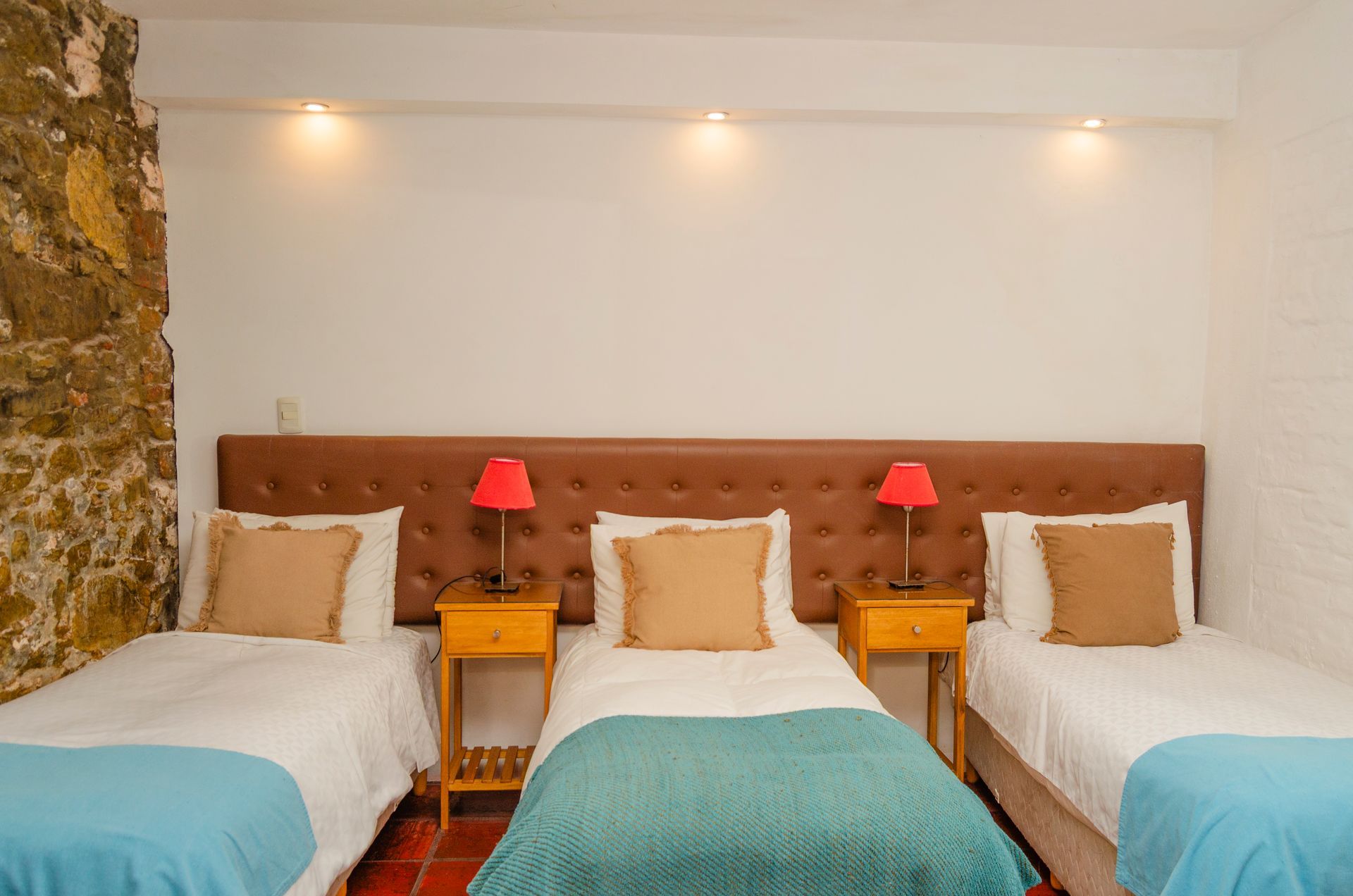 Tres camas con ropa de cama blanca, mantas color turquesa y almohadas color canela, apoyadas contra un cabecero marrón en una habitación con una pared de piedra.