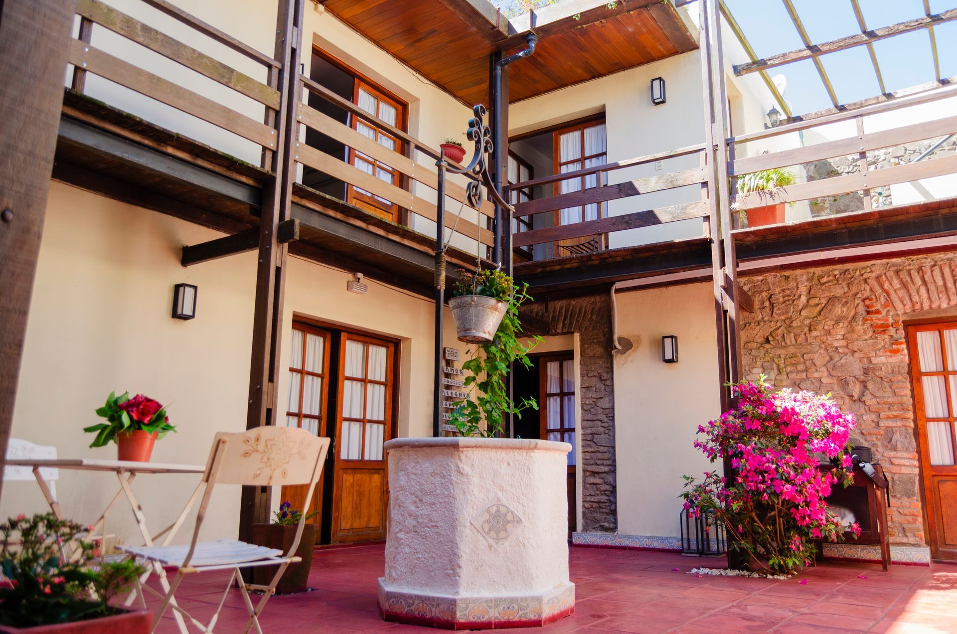 Un patio con suelo de baldosas rojas, un pozo de piedra, balcones de madera, paredes blancas y una mesita con plantas en macetas.