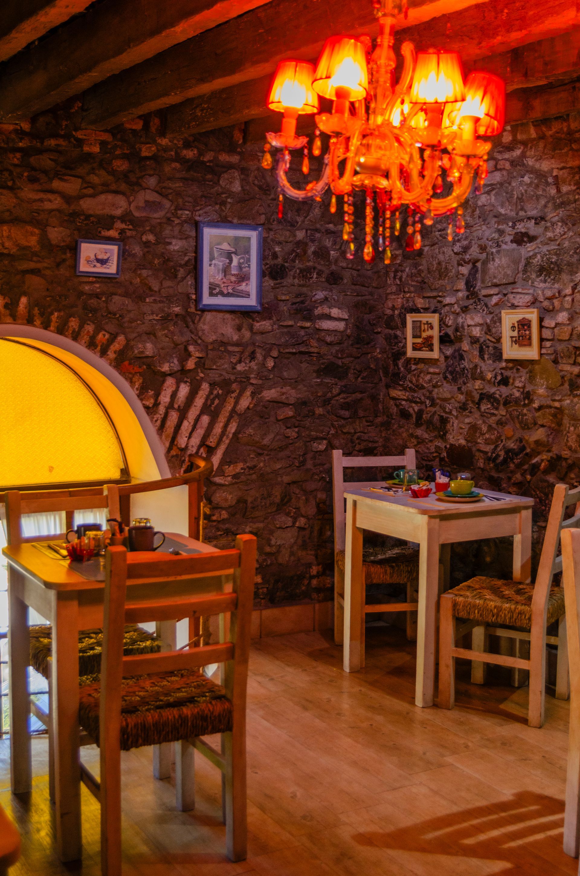 Un acogedor interior de restaurante con paredes de piedra, mesas de madera, sillas con asientos de mimbre y una cálida y brillante lámpara de araña en lo alto.