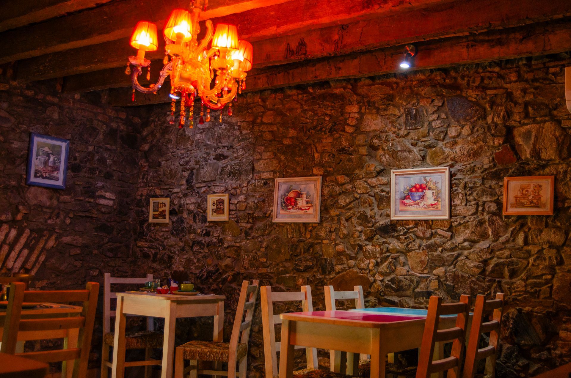 Un comedor rústico y con poca luz, con paredes de piedra, sillas y mesas de madera, y una lámpara de araña de color naranja vibrante en el techo.