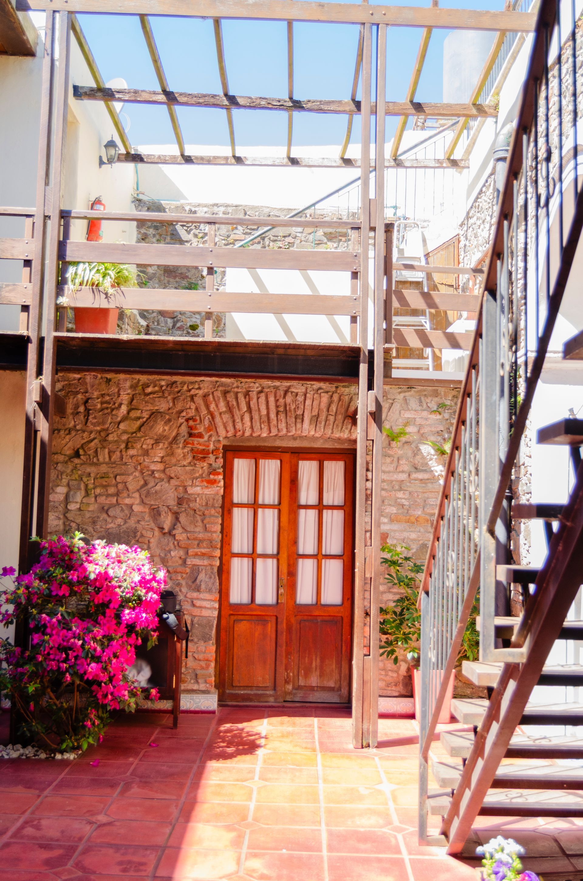 Un edificio rústico de piedra con una puerta de madera, una planta de flores de color rosa intenso y una escalera metálica exterior en un patio.