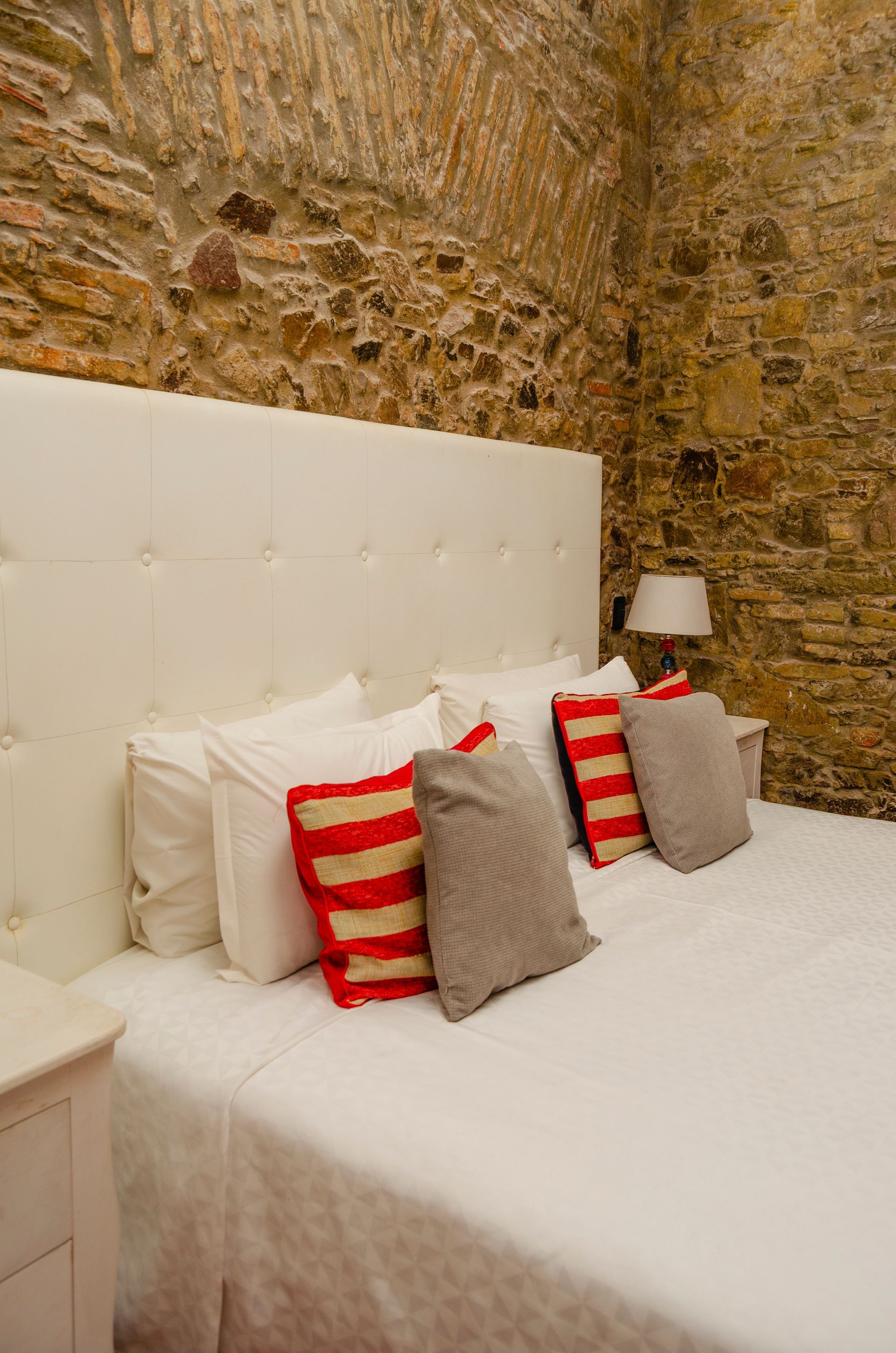 Una cama con cabecero blanco capitoné, sábanas blancas y cojines decorativos estampados contra una pared de piedra texturizada.