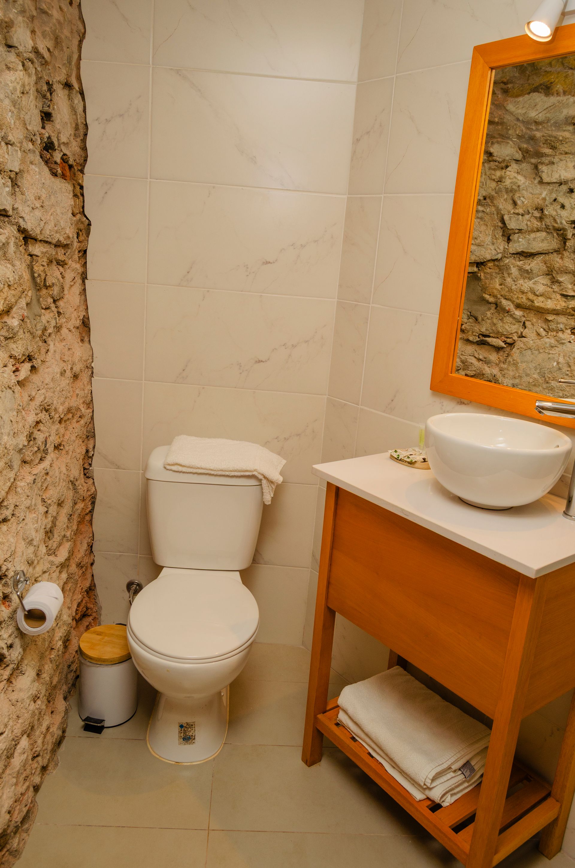 Un baño con inodoro, un tocador de madera con lavabo blanco sobre encimera, un espejo con marco de madera y una pared de piedra texturizada.