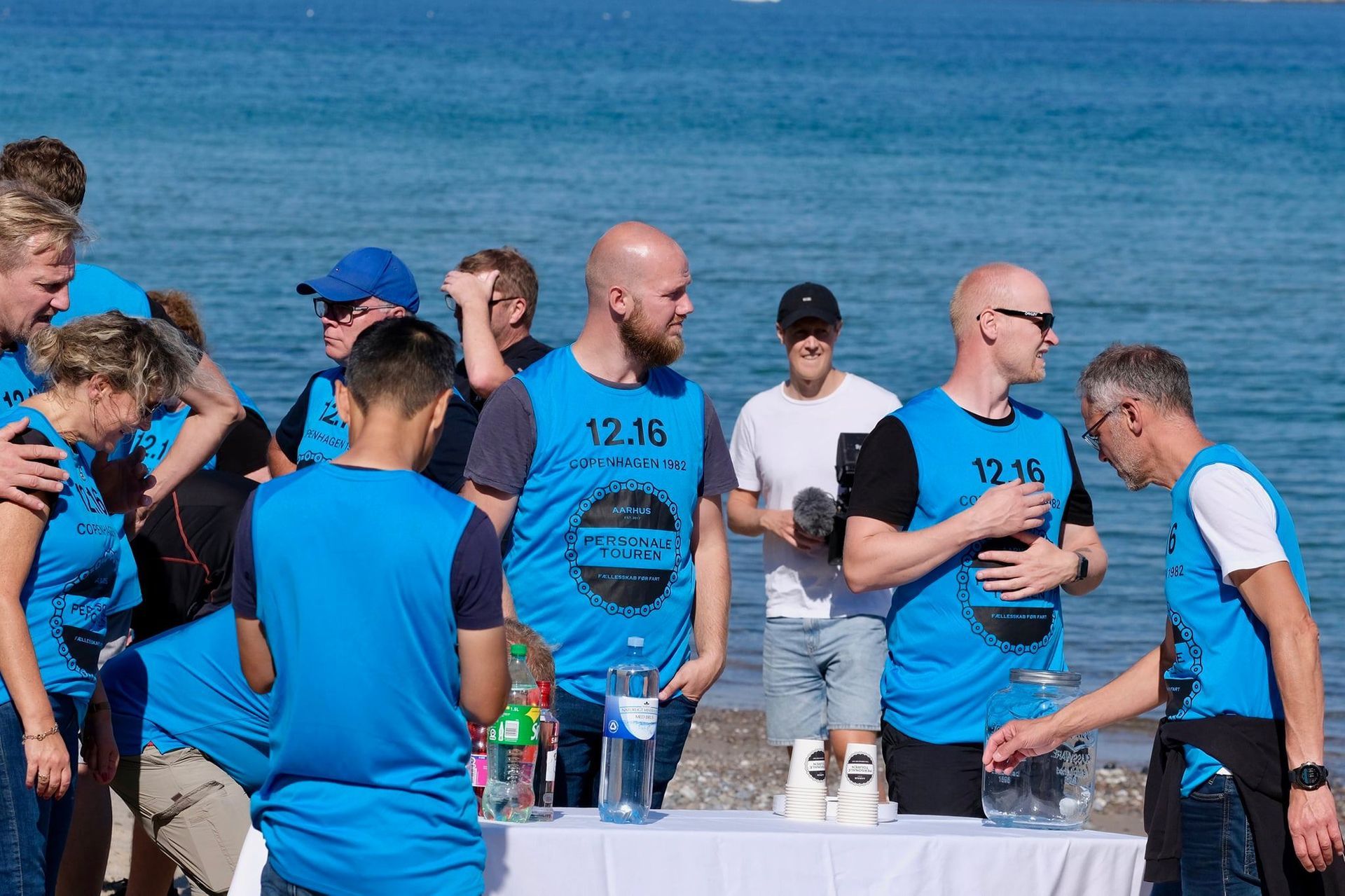 En gruppe mennesker i blå skjorter står omkring et bord på stranden.