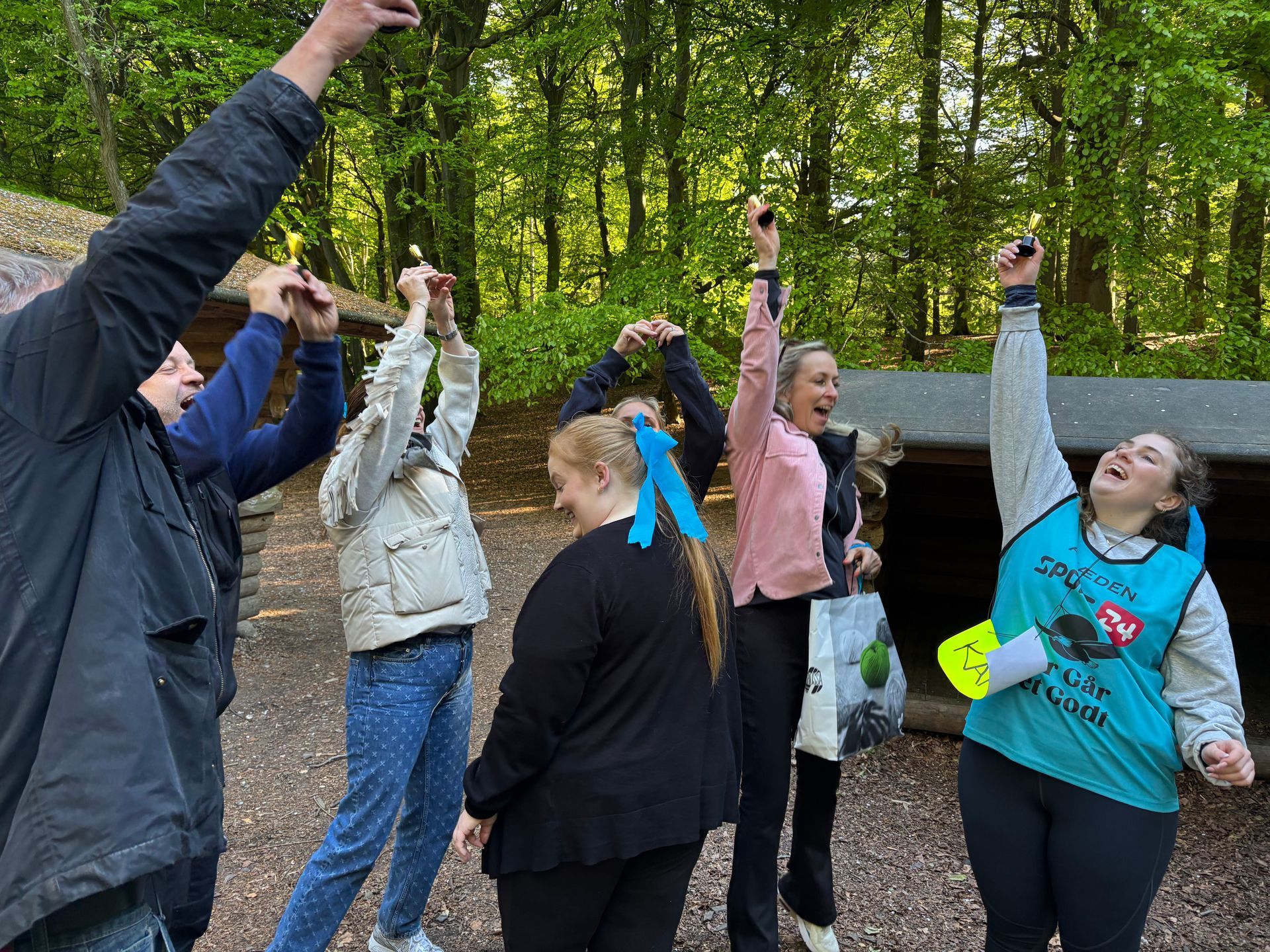 Gruppe af mennesker i en skov, der begejstret løfter armene. En har en blågrøn vest på, andre i afslappet tøj.